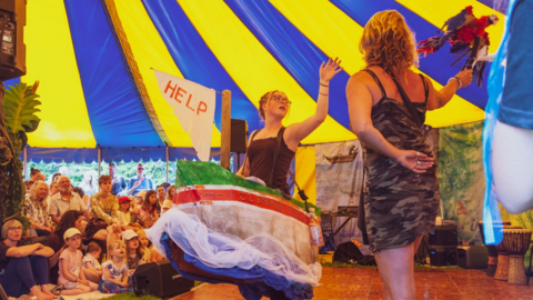 A blue and yellow-striped big top with two girls on stage. One is wearing a costume shaped like a boat with a large white flag saying 'help'. She is pointing towards a parrot puppet being flown by the other girl in the foreground. There are children sitting on the ground to the left of the stage and adults on chairs behind.