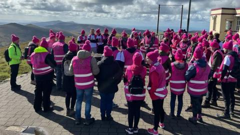 A large crowd of people wearing pink coats and pink woolly hats standing next to the cafe at the top of Snaefell, looking out at the hills on a sunny day.