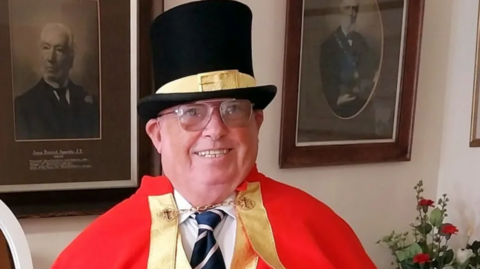 Man wearing a black top hat with a yellow buckled ribbon across the middle and is wearing a red cloak over a white shirt and blue stripy tie with a golden clasp fashioning the cloak.