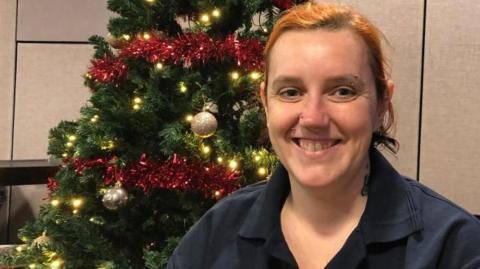 A woman is sitting in the BBC radio Suffolk studios with a Christmas tree  to one side and BBC branding the other. She is wearing a dark blue T-shirt and is smiling. She has her nose pierced and her eyebrow . 