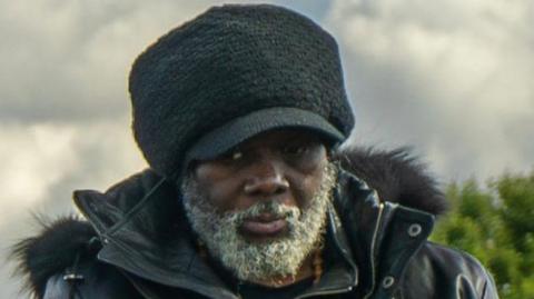 Franklin Francis in a black coat with a hood and a black hat. He has a grey beard.