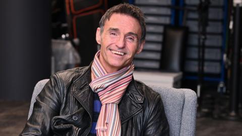 Marti Pellow sits in a grey chair smiling at the camera in the green room at BBC Radio 2. He wears a black leather jacket, a blue and black striped sweater and a multicoloured stripy scarf. A chair and a table are in the background.