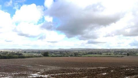 View Of The Up Mudford Urban Extension Site Somerset Council. There is a brown field of some acres and in the background, more fields and houses in the distance.