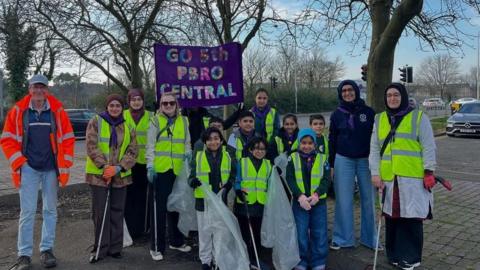 Adults and children in hi-vis jackets hold bin bags and litter picking tools. They are all smiling at the camera. There's a banner behind them that says "Go 5th Pbro Central".