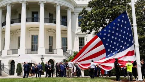 A US flag is held up by workers who are readying it up to be hung on a tall pole outside the White House as Trump and others look on.