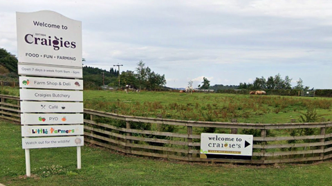Entrance sign for Craigies farm shop beside a fenced grassy field with grazing animals, listing facilities including a café, pick-your-own, butcher, and farm shop, under a cloudy sky.