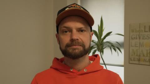 A man in a red hoodie wearing a black baseball cap. He is stood in a cream room with a plant and window in the background.