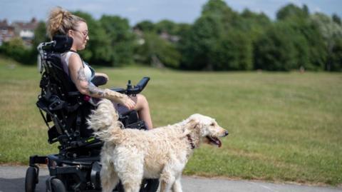 A woman with blonde hair, tattoos and a white top in a black wheelchair alongside a white dog on a footpath with grass alongside.