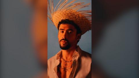 Bad Bunny has a black goatee and is wearing a straw hat and a cream shirt.