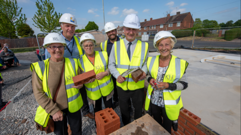 A group of six people wearing yellow high-visibility vests and standing in front of a block of bricks. Two women are holding a brick between them. There are houses and trees in the background. 