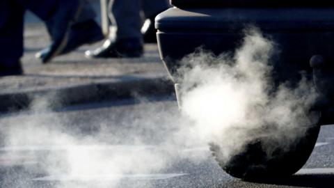 A small plume of exhaust fumes coming out of a car's exhaust pipe as it drives past some people walking on the pavement. Only the pedestrians' feet and legs are visible.