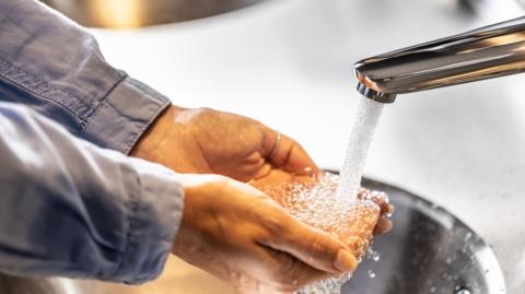 Water running out of a tap onto a person's hands which are cupped. You can only see the grey sleeves of their shirt.