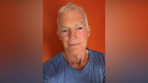 An older man with white hair. He is smiling at the camera and wearing a blue t-shirt.