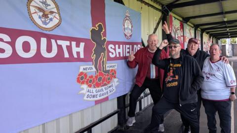 In a white, corrugated metal-backed stand, four supporters aged over 50 huddle together and raise fists in a joyous cheer. To their right hand side is a large South Shields flag in the clubs traditional claret and blue colours. 