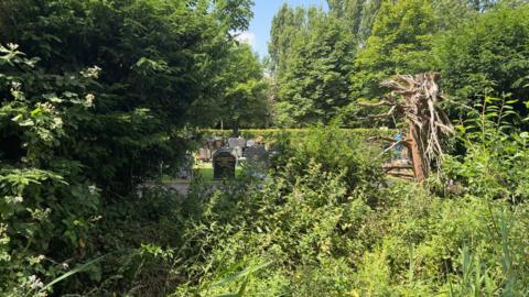 In the foreground of the photo there are lots of bushes and trees. Through the trees, there are a number of black and grey gravestones, which have flowers and different tributes on them. 