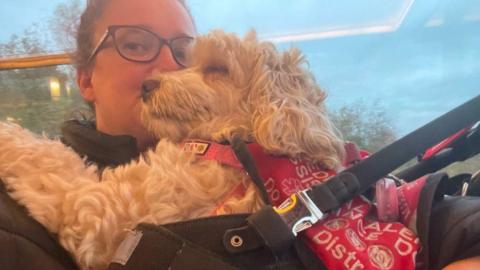 A woman with glasses and brown hair is cuddling her dog, of a cockapoo-type breed. The dog has a red jacket on that reads 'assistance dog. Do not distract.'