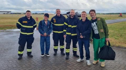 Six adults stand side by side on an airfield runway. Three of the adults are wearing dark firefighter uniforms with bright yellow reflective stripes and heavy black boots. One adult is dressed casually in a denim jacket over a hoodie, a baseball cap, loose blue jeans, and light trainers. Another adult is wearing a dark zip-up jacket, blue jeans, and white trainers. The final adult is wearing a green patterned jacket over a black T‑shirt with bold white text, bright green trousers, beige trainers, and is holding a large black bag. A helicopter is parked behind the group on a grassy area, with airport buildings in the distance under a cloudy sky.
