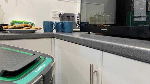 A grey and green bin labelled 'food waste' is in the foreground. Behind it there is a grey kitchen counter with cookies, bread and three mugs arranged in front of a toaster and next to a microwave.