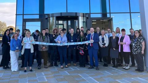 A group of people stood outside a glass building. They are cutting a ribbon.