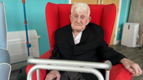 Old man with short grey hair in black blazer and white shirt sat in red chair 