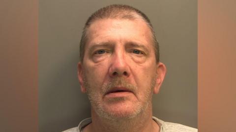 A police mugshot of a man with short brown hair and moustache and grey beard stubble. He is wearing a grey top and looking directly into the camera against a dark wall.