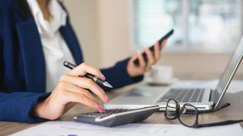 Close up image of a professional woman sitting at a computer making calculations and looking at her smartphone.  She is wearing a blue blazer and an open neck white blouse. (Stock image) 