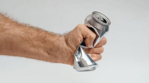 Male hand squeezing and crushing empty aluminum beverage can on gray background. Symbolic gesture expressing stress, frustration, anger or emotional pressure.