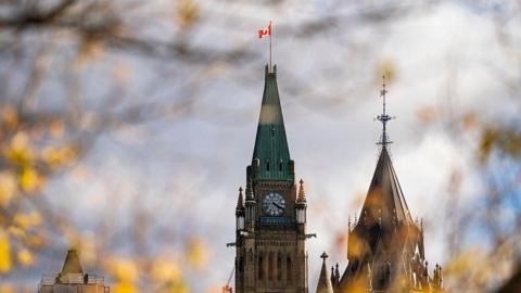Fall leaves surround the Peace Tower on Parliament Hill. It has a green roof and features a large clock. At the top of its roof is a Canadian flag. 