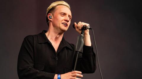 A man with blond hair, standing onstage with a microphone