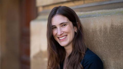 Katherine Rundell smiling leaning against a building. She has long dark hair and is wearing a navy coloured top.