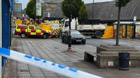 A street which has been cordoned off with police tape and there are several vehicles within the cordon, including trucks and a fire service vehicle. 