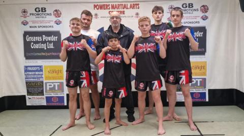 Six martial arts athletes and coach pose with raised fists in a gym, wearing matching ‘Powers-Mac’ uniforms in front of sponsor banners.