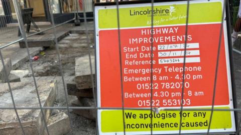 A metal sign displays text to warn residents of repairs taking place between July and September. The foundations of the street can be seen in the background, with paving slabs removed.