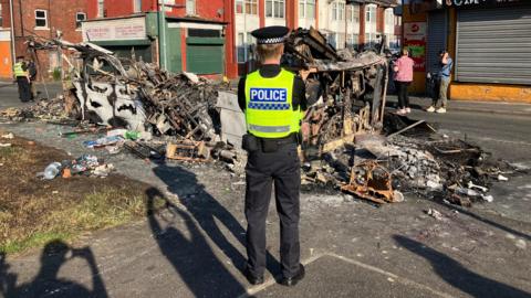 A police officer guards the scene of a burned out bus