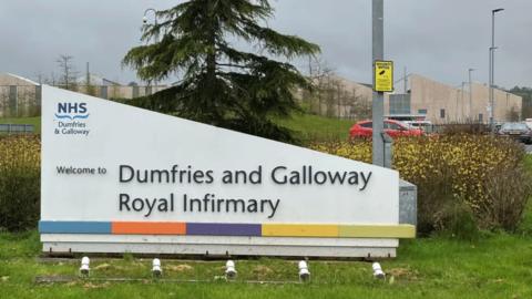 A sign for Dumfries and Galloway Royal Infirmary in front of a tree and a lamp post and a car park with wooden hospital buildings in the distance