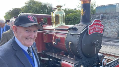 Raymond Gawne, a man wearing a suit and a baseball cap has a bright smile as he stands in front of a red steam train.