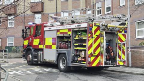 A fire engine is parked on an area of road marked NO PARKING. Behind it is a low block of residential flats with a brick exterior and lots of windows. A piece of cordon tape is tied to a lamppost and extends out of the picture.