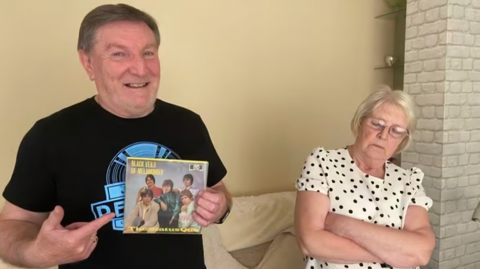 Kevin Turner alongside his partner Janet. He is holding a record while wearing a black T-shirt with a blue logo while his partner feigns sleep by resting her head against a wall