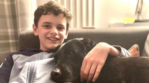 A boy in a light-coloured hoody with a dark stripe on the arms and shoulders is sitting on a sofa and hugging a dog. The boy is smiling and looking at the camera