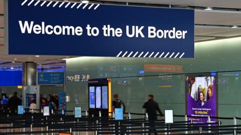 A general photo of UK border controls at an airport.  There is a sign saying "welcome to the UK border" and people are queuing in cordoned off lines for documentation checks