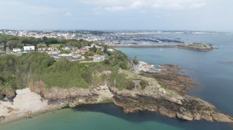 Fort George from the sky - A cliff face from the sky, with leafy green growth on the side of the cliff face and some houses atop it. 