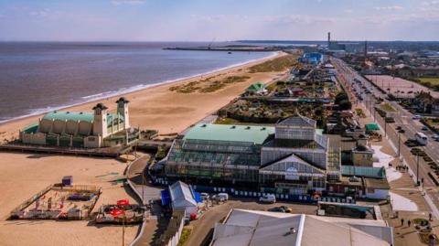 An aerial view of Great Yarmouth seafront, showing the golden sandy beach and calm sea to the left and, in the centre of the image, a large glasshouse known as Winter Gardens. To the right, a road stretches out of sight, parallel to the sea. In the distance is the blue structure of the rollercoaster at the Pleasure Beach.