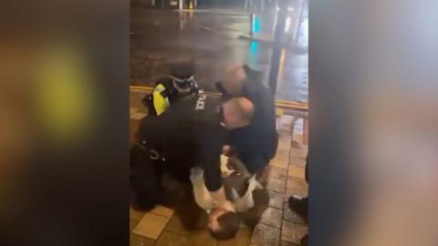 Colin Allan, in a cream blazer, is being held face down on a pavement by three police officers, one of which has his hand on the back of Mr Allan's neck
