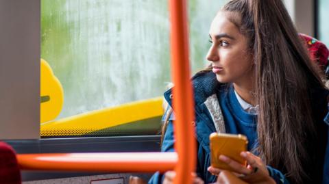 Teenage girl wearing a ponytail and a navy blue parka coat sits on the bus while holding her smartphone in her left hand and gazing out the window