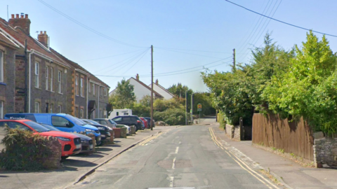 A google maps street view of Poplar Road in Warmley. It is a residential street with houses on one side. There are many vehicles parked on driveways in front of the houses. The other side of the road there is a fence and hedges.