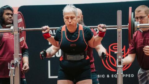 Dee Atkins-Greig in a powerlifting competition. The barbell is on her shoulders, her face shows effort.