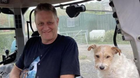 Ian Pullen sits in a tractor with Biskit the Parson's terrier.  He is wearing a black T-shirt and shorts, Biskit has a white coat