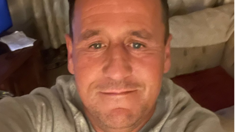 A selfie image showing a man in his 40s with receding brown hair. He is in a living room with a sofa and the corner of a TV visible. He is wearing a light grey top.