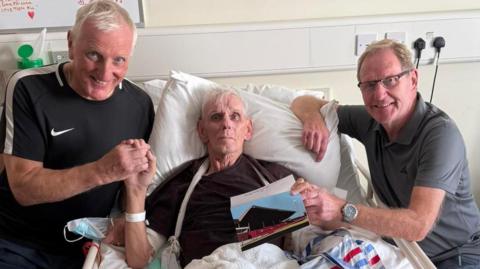 A man, Wayne Ryalls, in a hospice bed. At either side holding his hands are Ronnie Moore and John Breckin