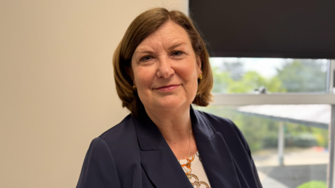 Wendy Reid has short straight brown hair. She is wearing a dark blue suit jacket over a patterned top. She is standing in an office in front of a window.